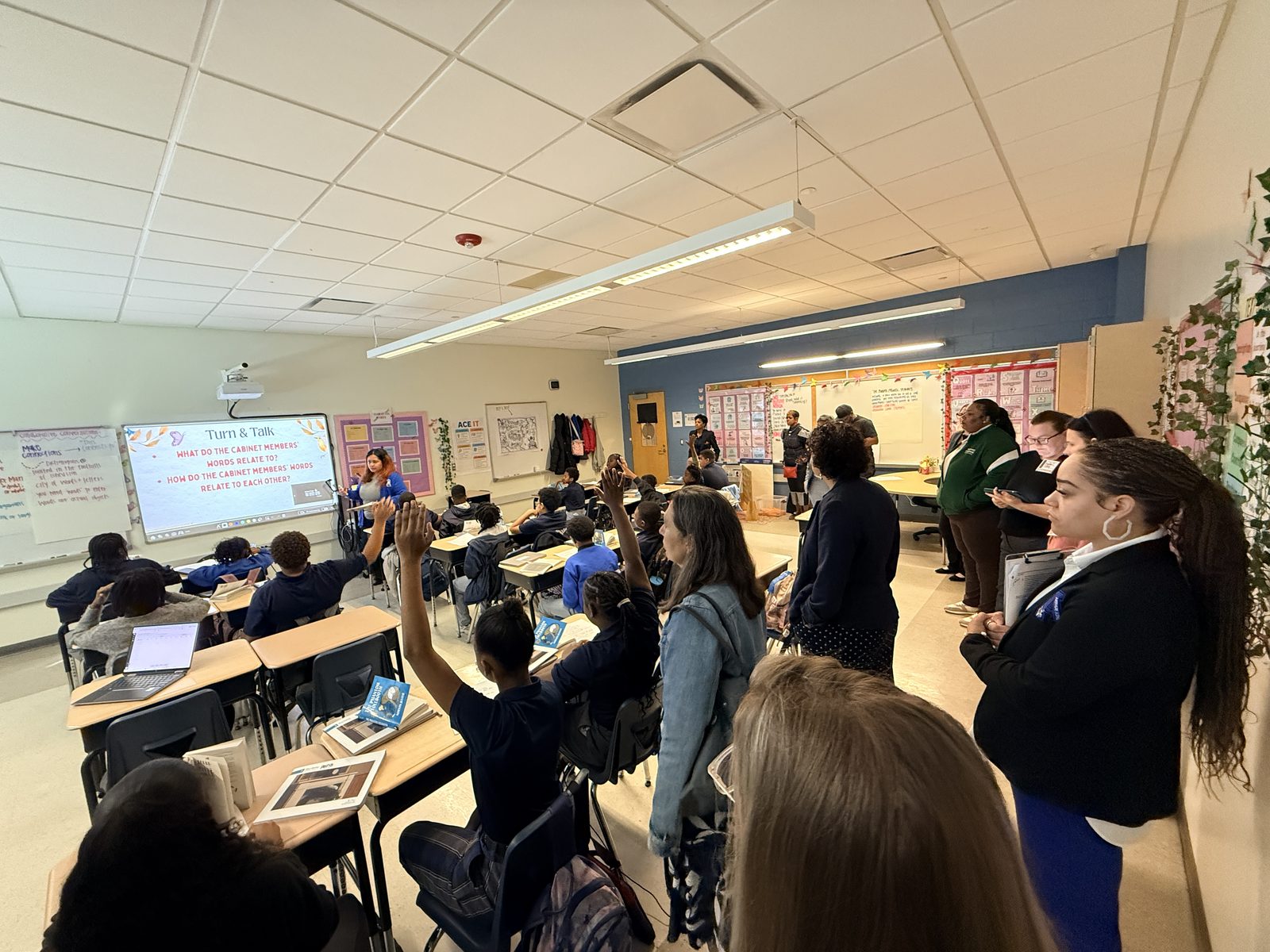 Students raise their hands during a lesson while visitors observe